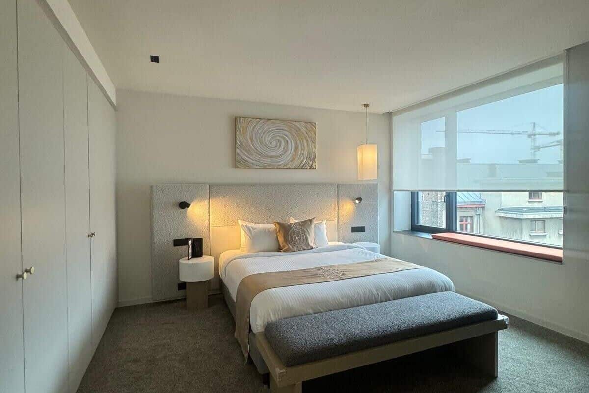 Une chambre d’hôtel moderne dotée d’un grand lit, de tables de chevet, d’une tête de lit avec un luminaire intégré, d’une fenêtre donnant sur des immeubles et d’un sol recouvert de moquette.
