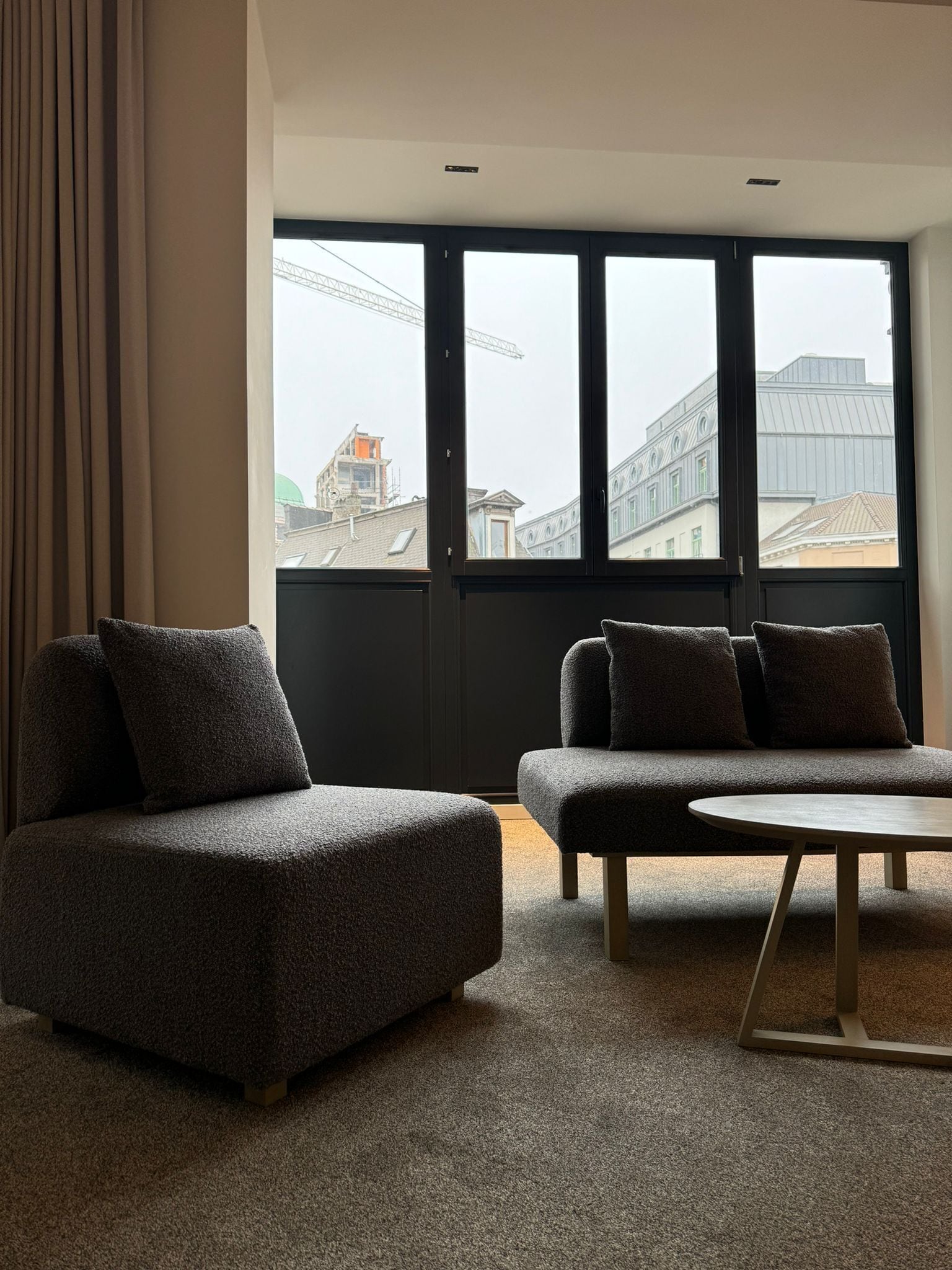 A modern living room with a sofa and ottoman, large windows with a view of buildings and a crane, and a carpeted floor.
