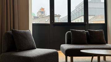 A modern living room with a sofa and ottoman, large windows with a view of buildings and a crane, and a carpeted floor.