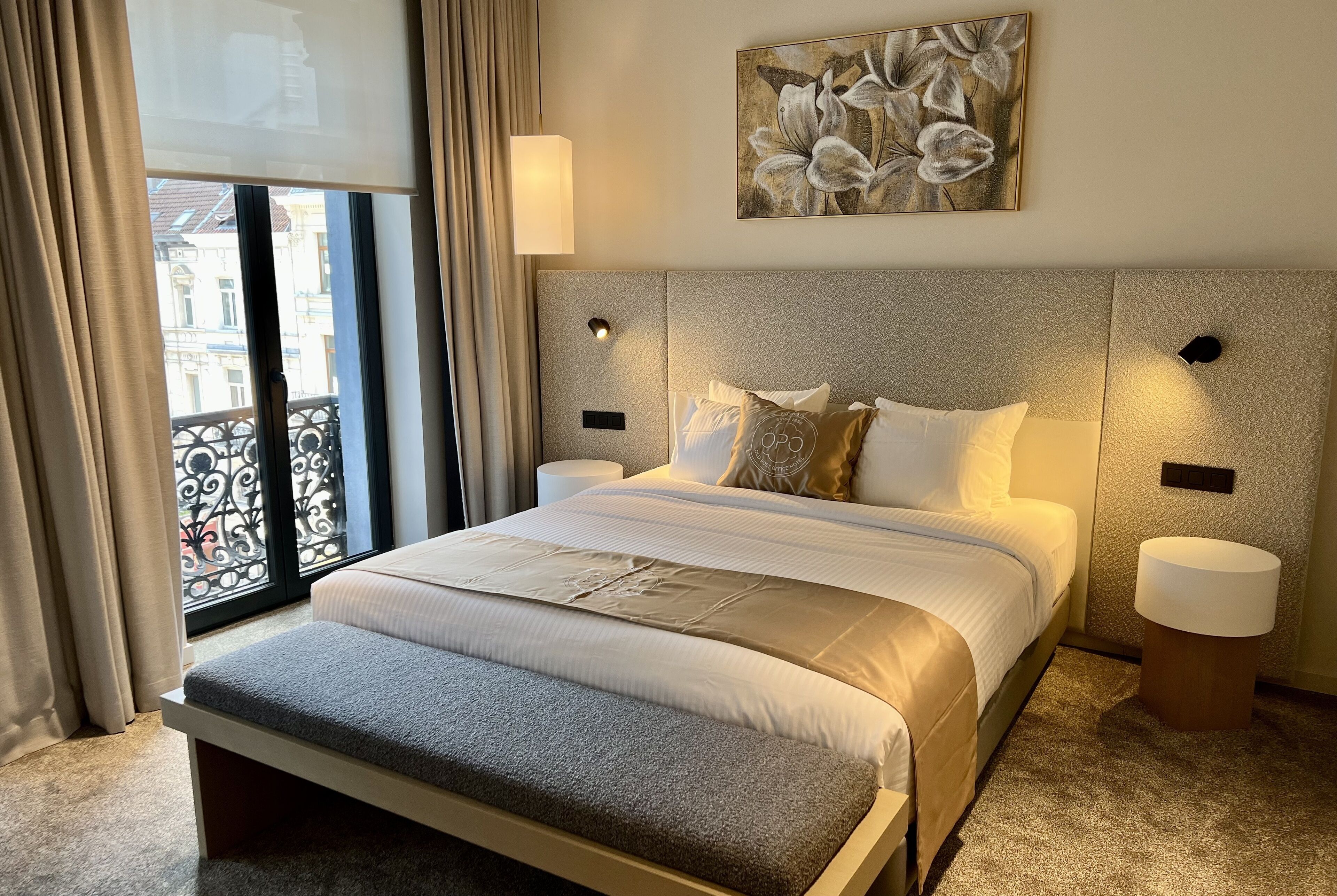 Une chambre d’hôtel bien aménagée, avec un grand lit, deux tables de chevet, une tête de lit agrémentée d’appliques murales et un balcon avec vue.
