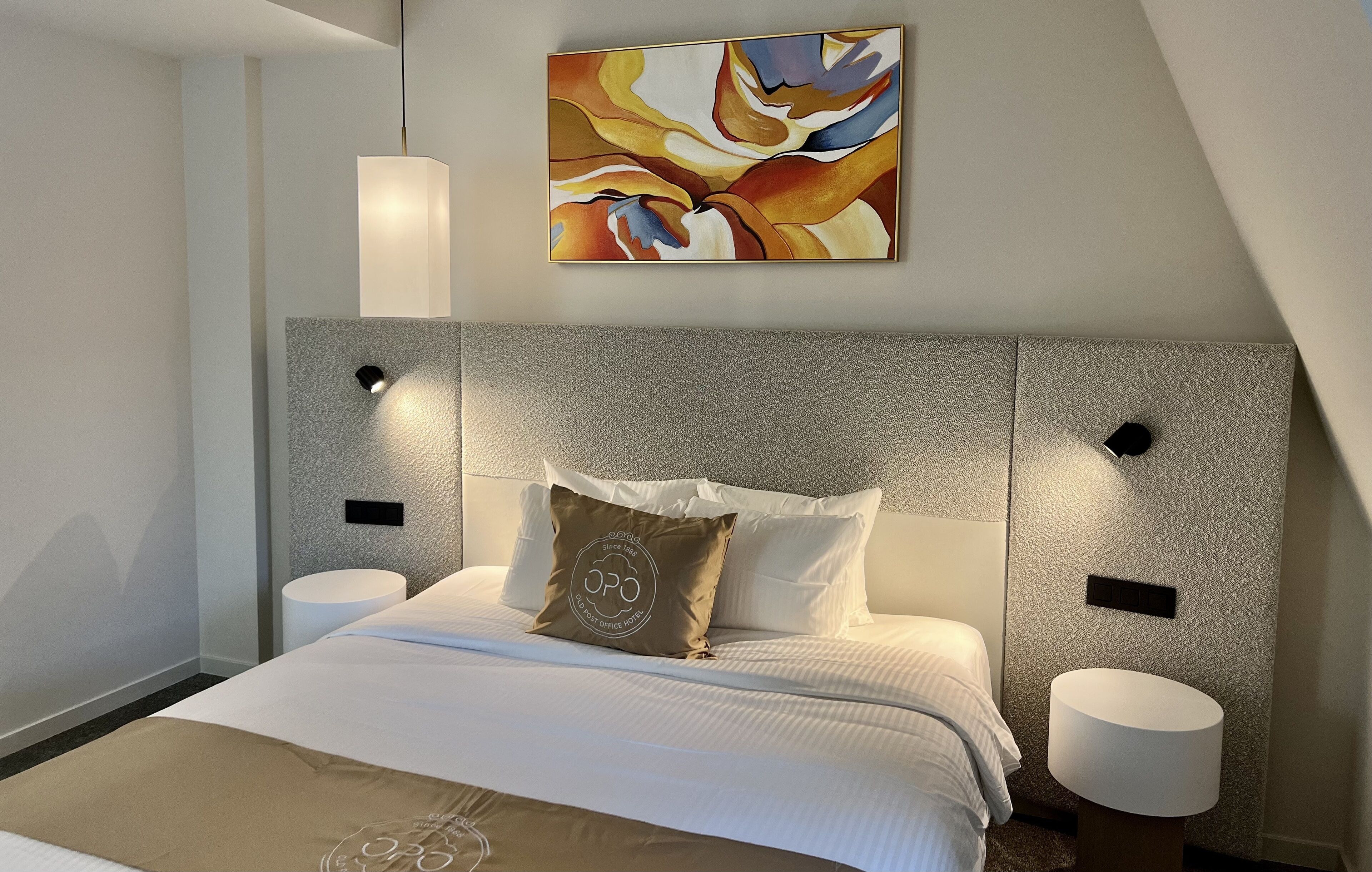 A modern hotel room with a large bed, a textured headboard, and a contemporary abstract painting above the bed.