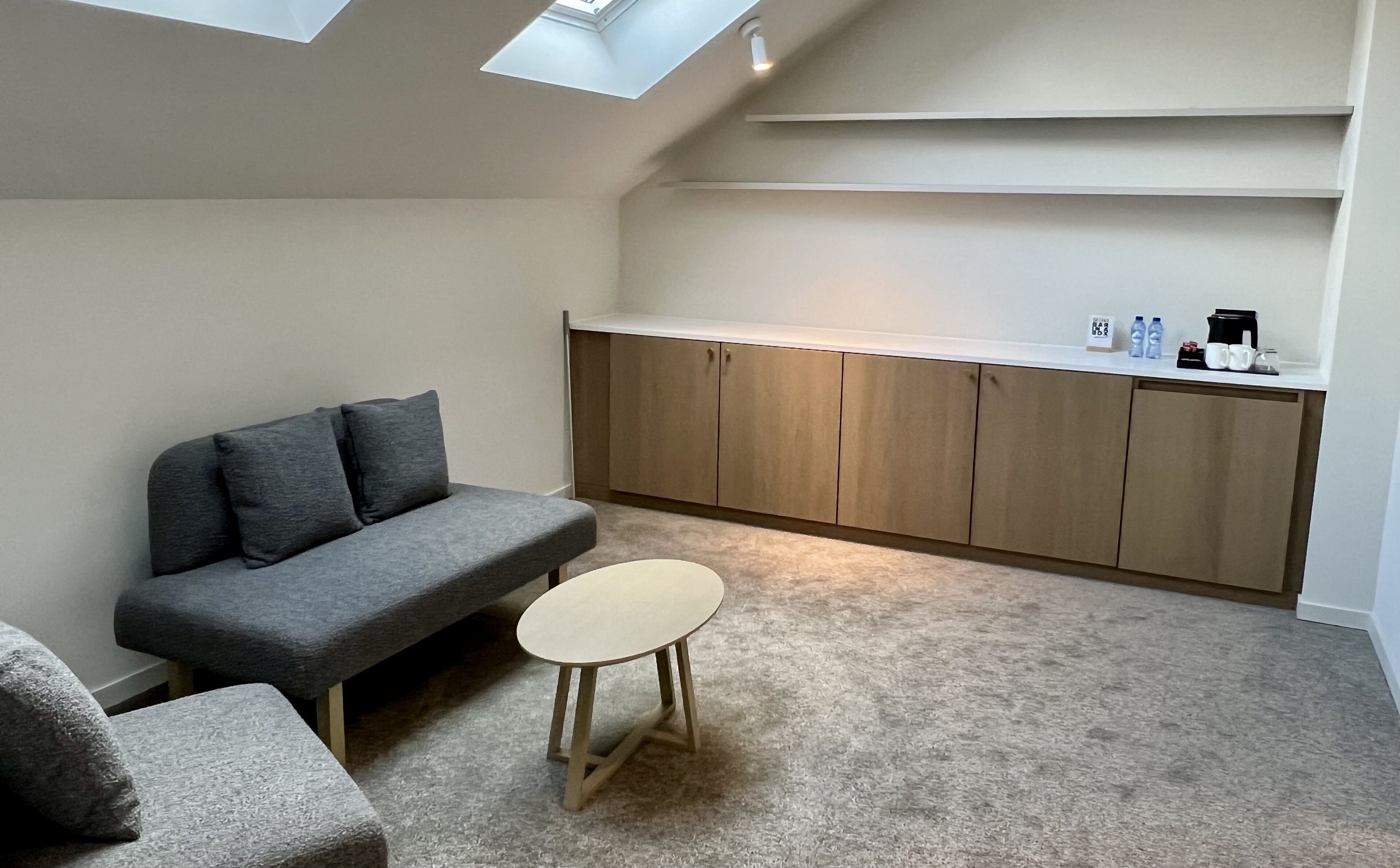 Un espacio habitable pequeño y funcional con un sofá gris, una mesita redonda y una zona de kitchenette con armarios y encimera.