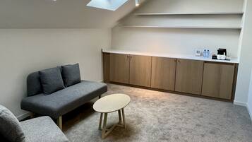 Un espacio habitable pequeño y funcional con un sofá gris, una mesita redonda y una zona de kitchenette con armarios y encimera.
