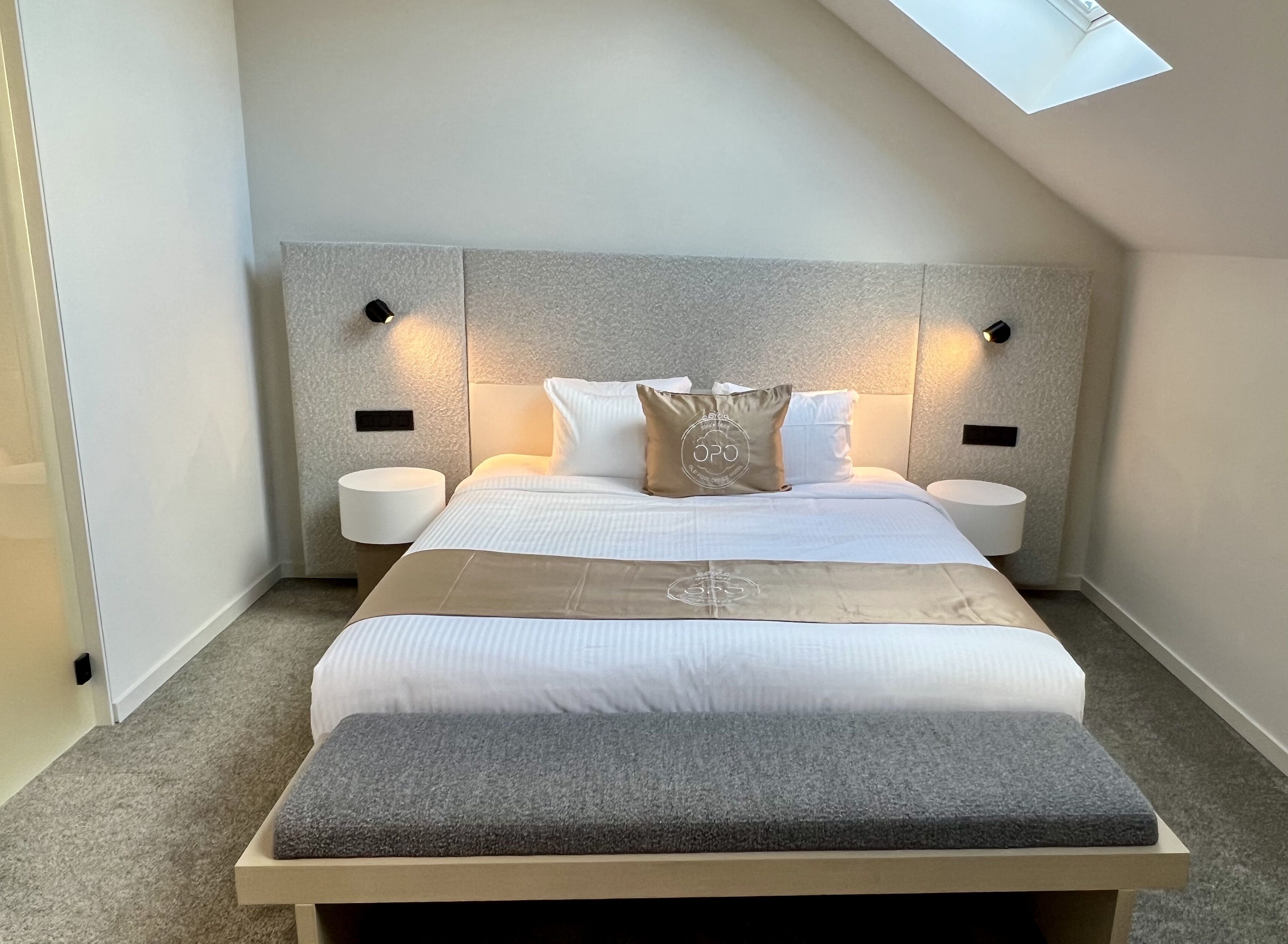 Ein modernes Schlafzimmer mit schrägem Dach, einem großen Bett mit grauem Sitzbank, zwei Nachttischen und einem Oberlicht.