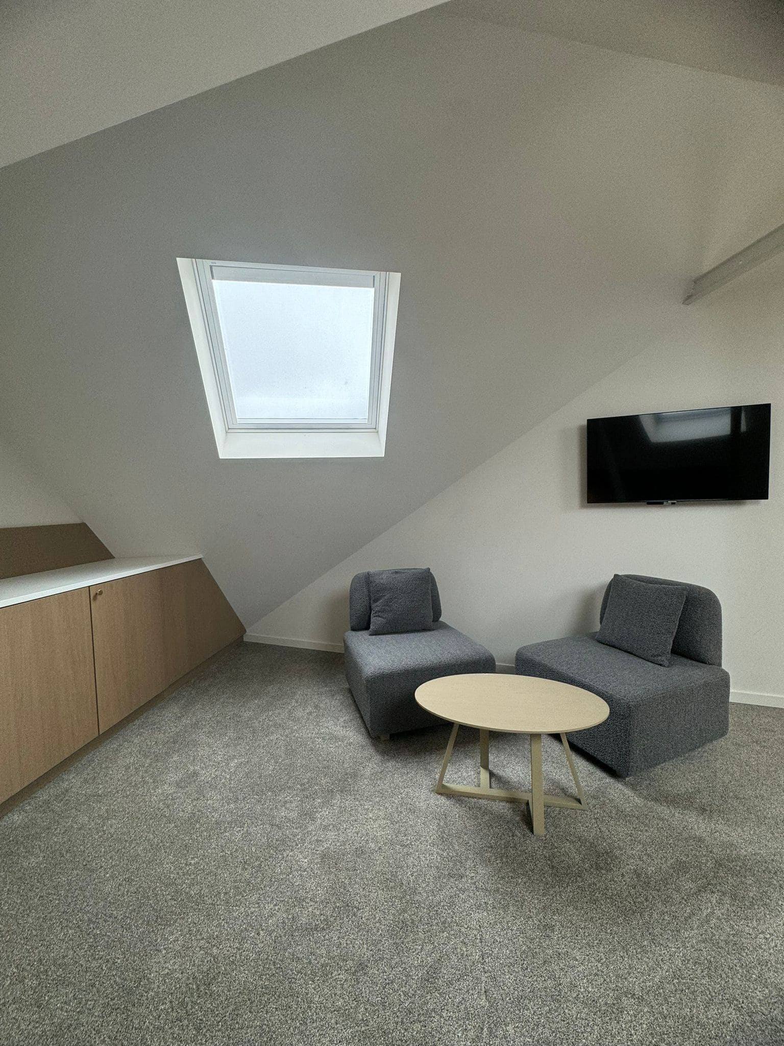 A modern loft bedroom with a sloped ceiling, a skylight, a flat-screen TV mounted on the wall, and a small round table.
