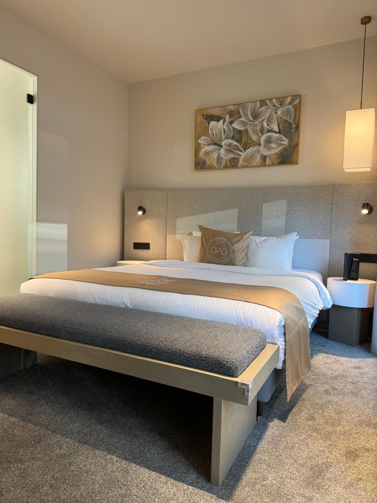 Ein modernes Schlafzimmer mit einem großen Bett, einem an der Wand befestigten Kopfteil mit integrierter Beleuchtung, einem Nachttisch mit Fernbedienung und einem floralen Kunstwerk.