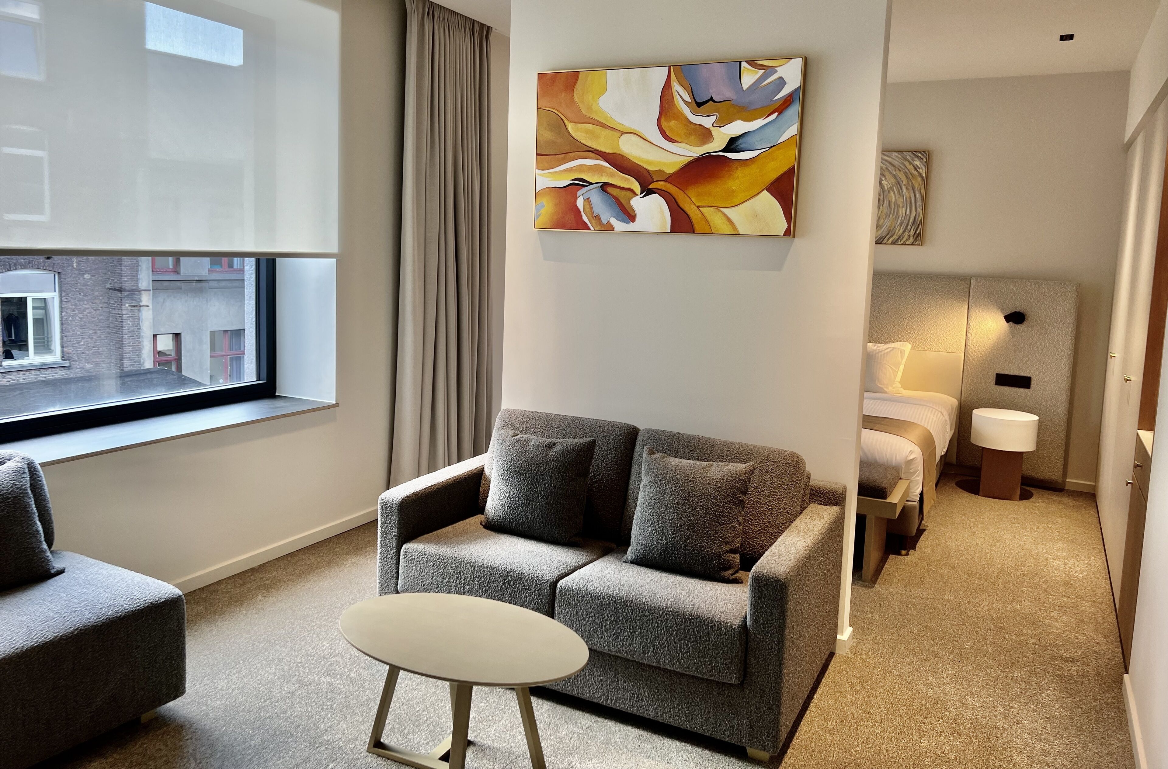 Quarto de hotel moderno com sofá cinza, uma mesinha redonda, cama com cabeceira branca e uma grande pintura abstrata na parede.