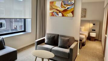 Quarto de hotel moderno com sofá cinza, uma mesinha redonda, cama com cabeceira branca e uma grande pintura abstrata na parede.
