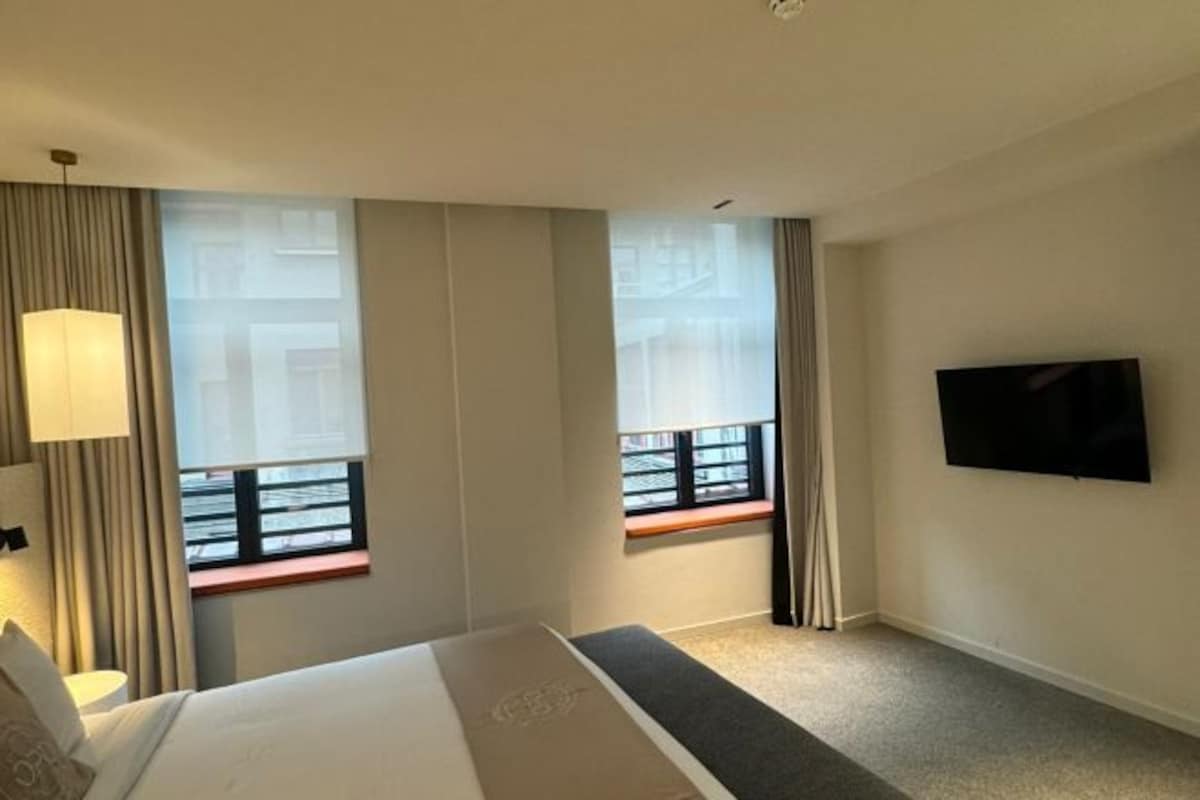 A hotel room with a bed, a TV mounted on the wall, and windows with curtains.