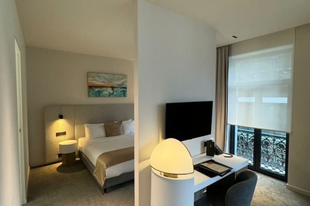 A hotel room with a bed, bedside table, lamp, TV, and a desk with a chair.