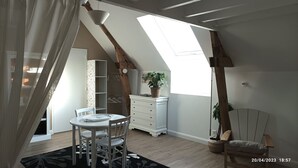 A room with a sloped ceiling, a dining table with chairs, a white dresser, a plant on the dresser, a plant in a pot, and a chair with a cushion. - Grand Studio 4 Couchages à 450m de la mer (Hermanville-sur-Mer)