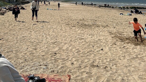 Una playa con gente caminando, algunas mantas sobre la arena y edificios al fondo.