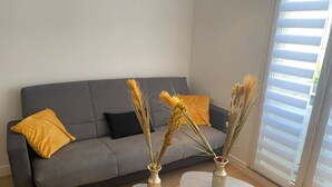 Una sala moderna con un sofá gris, dos mesitas redondas con jarrones y una ventana con persianas.