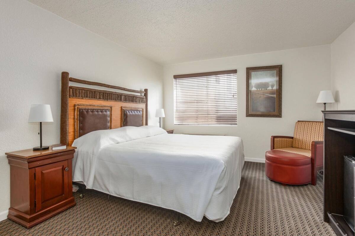 A hotel room with a large bed, a wooden headboard, a nightstand with a lamp, a chair, a small ottoman, and a television.