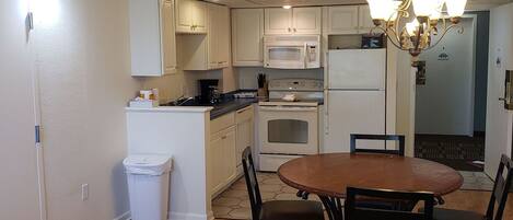 A compact kitchen with white cabinets, a microwave, an oven, and a refrigerator. There's a dining table with four chairs and a chandelier above.