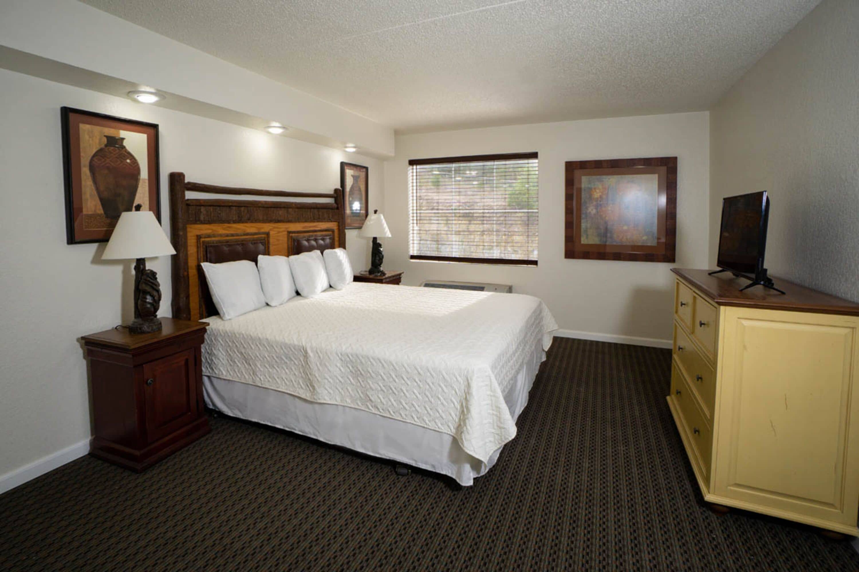 A hotel room with a large bed, a wooden headboard, a television on a stand, a dresser, and two framed pictures on the wall.
