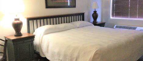 A neatly made bed with a white comforter, a bedside table with a lamp, and a window with blinds.