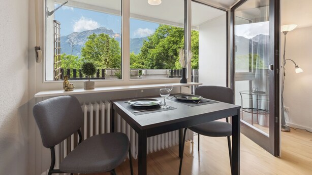A modern dining area with a table set for two, large windows offering mountain views, and a glass door leading to an outdoor balcony. - Apartment BERGBLICK (Bad Reichenhall)