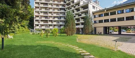 A multi-story residential building with balconies, a grassy courtyard, and a paved pathway leading to a parking area.