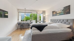 A modern bedroom with a large bed, a dining area, and a view of greenery through large windows. - Apartment BERGBLICK (Bad Reichenhall)