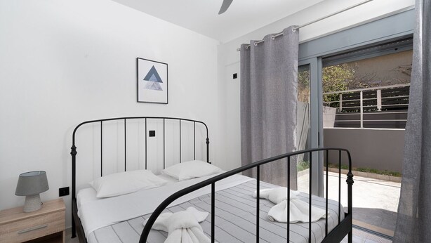 A bedroom with a bed, a nightstand, a ceiling fan, a window with curtains, and a view of the outside. - MTC-AN Apartment by the sea and Airport (Markopoulo Mesogaias)