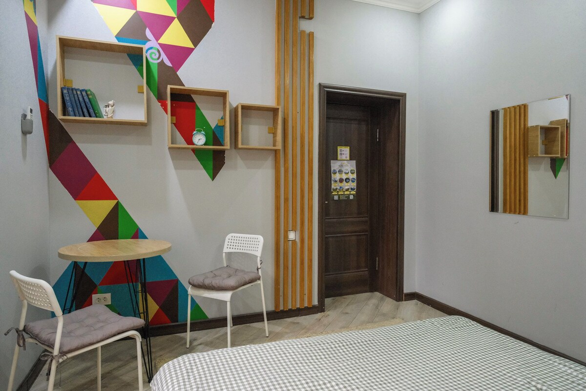 A room with a checkered floor, two white chairs, a small round table, a wooden door, and a colorful geometric wall design.