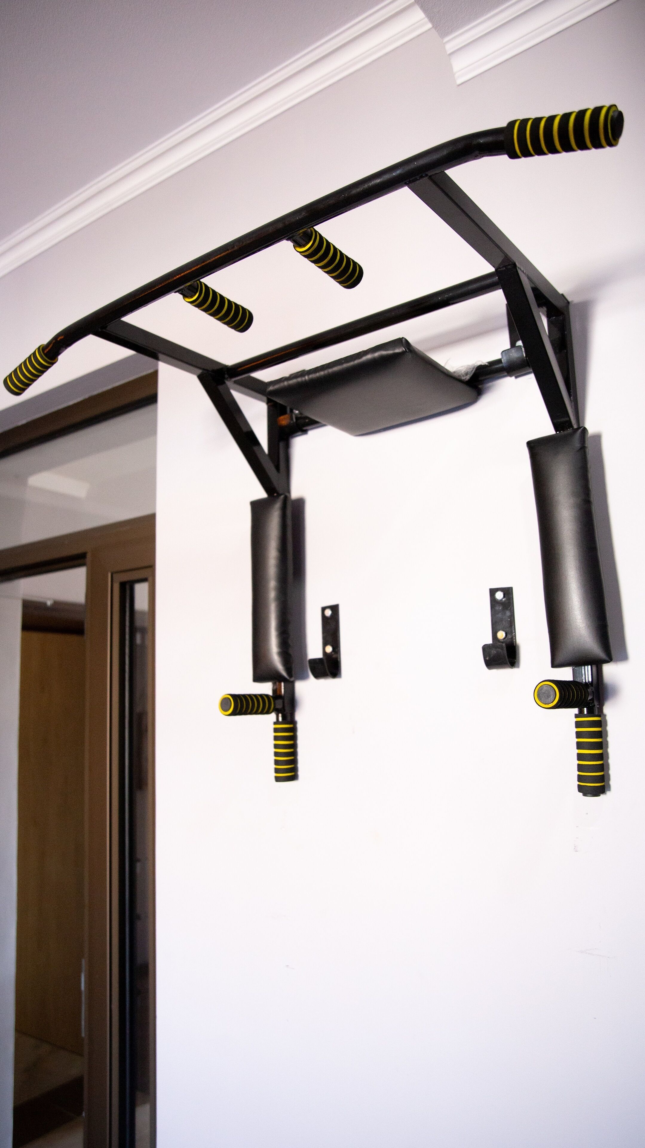 A wall-mounted pull-up bar with yellow and black grips, installed in a room with white walls and a wooden door.