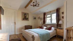 A bedroom with a large bed, a wooden dresser, a nightstand, a lamp, a window with curtains, and a painting on the wall. - Secluded stables with parking in grounds of home in the heart of the new forest  (Burley, Nr Ringwood)