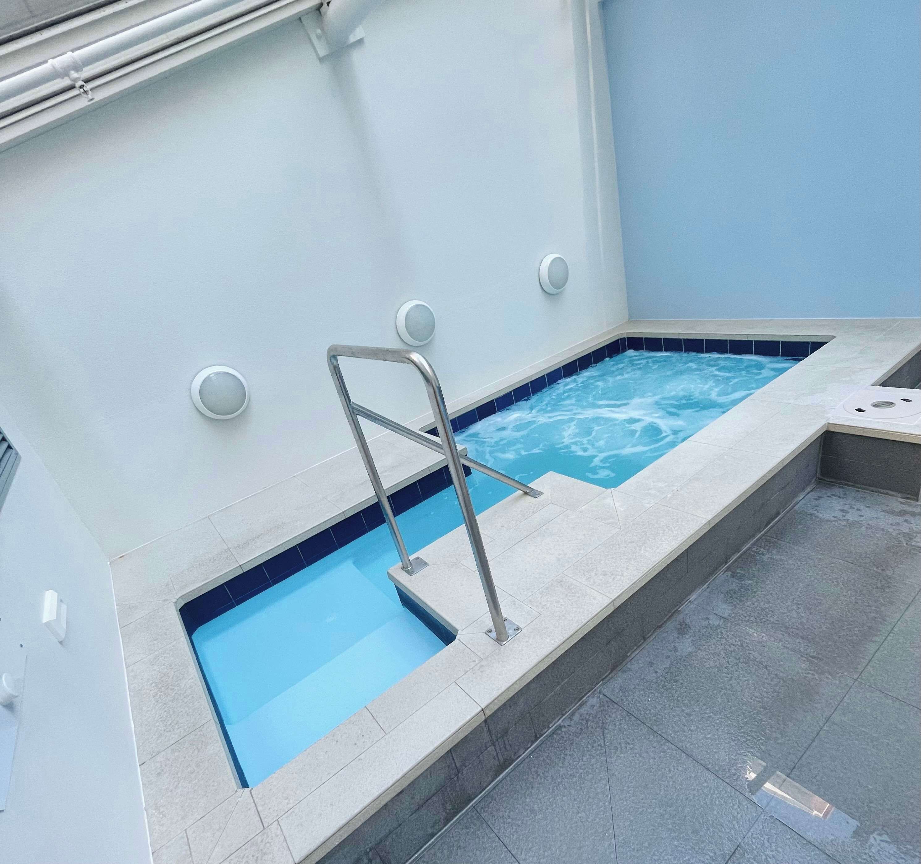 Une petite piscine intérieure avec un escalier en métal, un élément décoratif aquatique bleu et un sol carrelé.