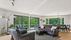 Un salon spacieux avec un canapé en cuir, une table basse et un coin repas comprenant une table et des chaises. De grandes fenêtres offrent une vue sur la verdure environnante.