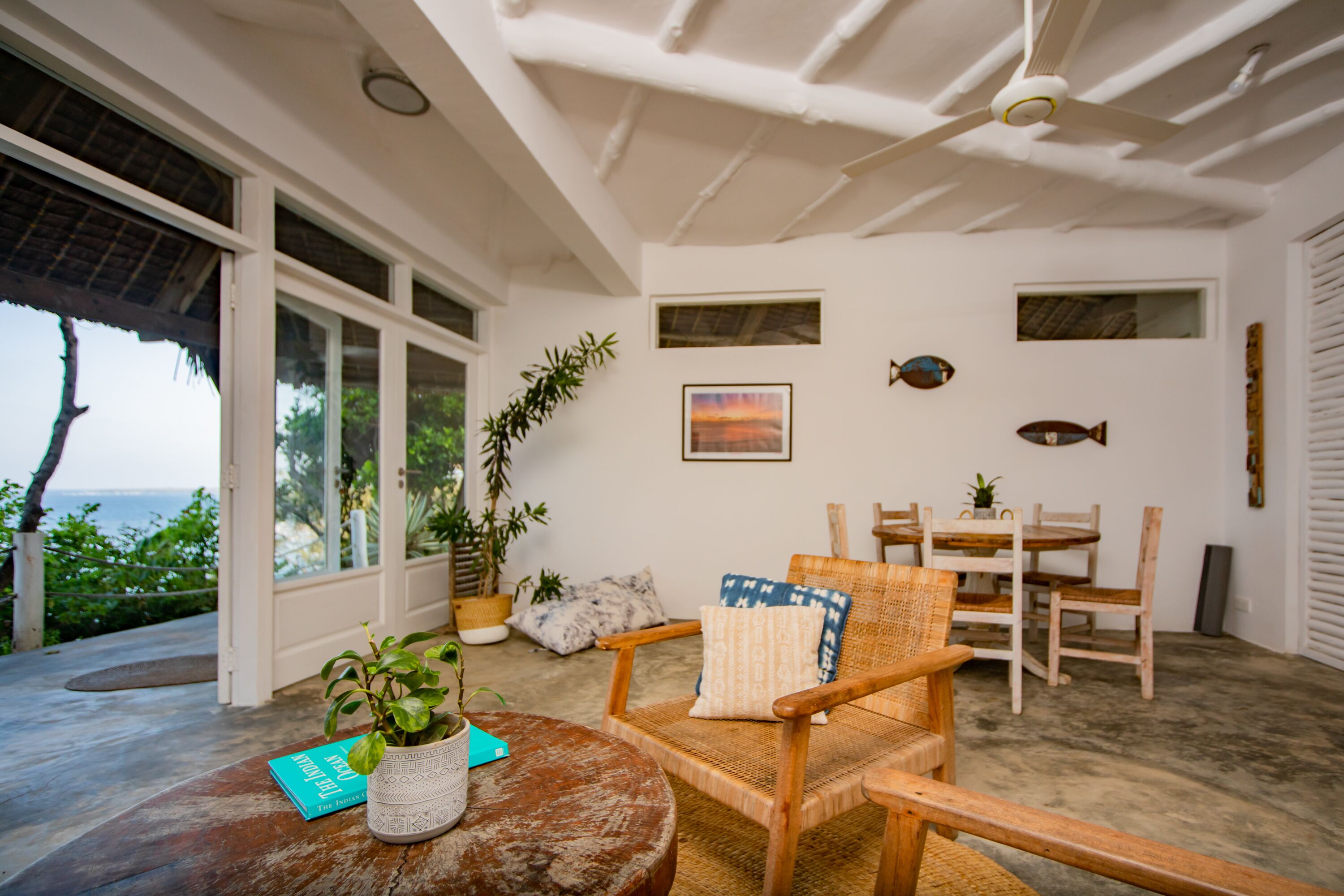 A room with a large window, a dining table, chairs, a wooden chair with a cushion, a potted plant, and a ceiling fan.