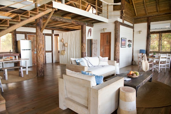 A spacious room with a wooden floor, a bed with white bedding, a wooden bench, a dining table with chairs, and a large window.