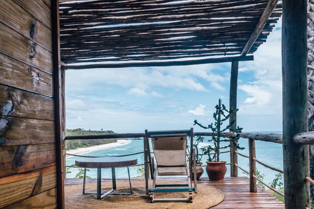A wooden deck with a view of the ocean, a chair, a small table, and a potted plant.