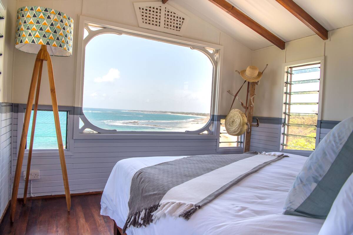 A bedroom with a large window overlooking the beach, a bed with white linens, a wooden floor, and a patterned floor lamp.