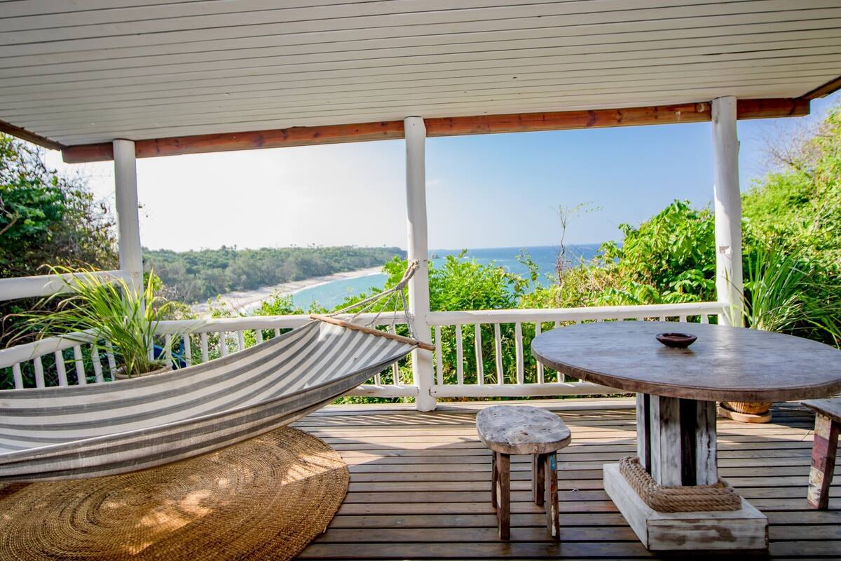 A wooden deck with a hammock, a round table, and a bench, overlooking a beach and ocean.