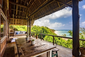 Une terrasse en bois, surmontée d’un toit de chaume, qui offre une vue sur une plage et l’océan.