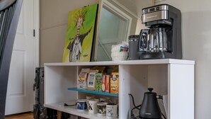 Eine Küche mit Kaffeemaschine, Wasserkocher und einem Regal mit Kapseln und Tassen.