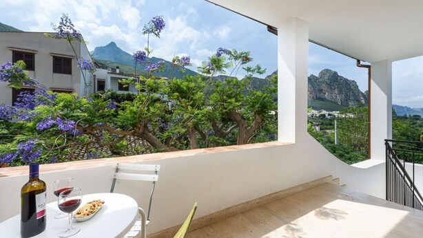A balcony with a table set for two, a bottle of wine, and a plate of food. The balcony overlooks a lush garden with purple flowers and a mountain in the background.