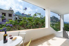 A balcony with a table set for two, a bottle of wine, and a plate of food. The balcony overlooks a lush garden with purple flowers and a mountain in the background. - I Tre Golfi - Casa ChiaraMarina (San Vito Lo Capo)