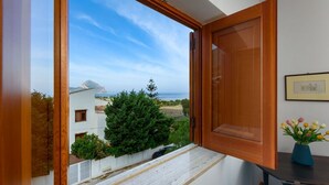 A room with a large window offering a view of a coastal landscape, a wooden door, and a small table with a vase of flowers.