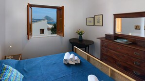 A bedroom with a bed, a dresser, a small table with a vase, and a window with a view of a mountain and buildings.