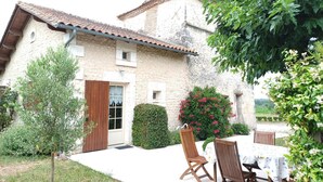A rustic stone building with a tiled roof, a wooden door, and a small window. There is a garden with a table and chairs, and a variety of plants and flowers. - Charming Cottage with WiFi and Pets Allowed (Saint-Romain)