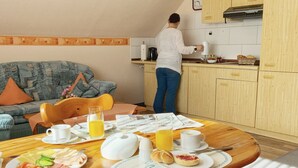 Una sala de estar acogedora con un sofá, una mesa de comedor de madera dispuesta para el desayuno y una cocina con armarios de madera y una persona preparando alimentos.