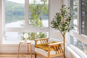 A room with a large window, a white chair with a yellow throw, a small table with a white object, and a potted plant.