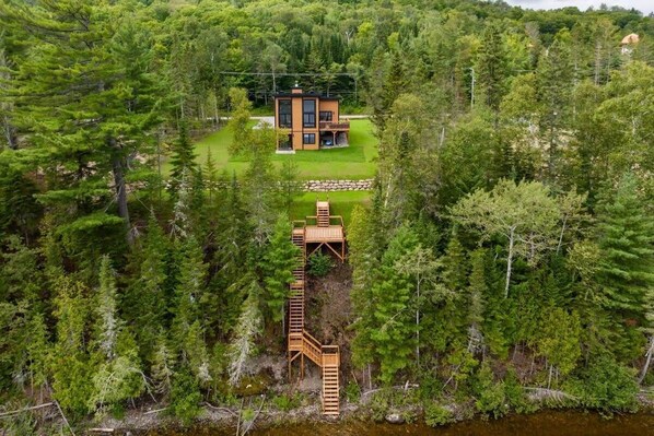 A modern two-story cabin surrounded by dense forest, with a wooden deck and a staircase leading to a small platform.