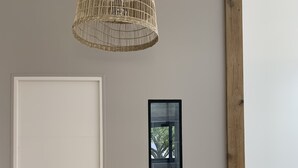 Una habitación con una puerta blanca, una lámpara colgante de fibra natural, un cuadro enmarcado y un pilar de madera.