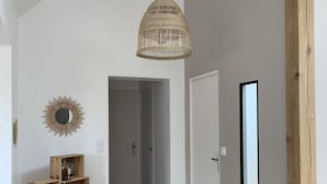 Un pasillo con suelo de madera, una lámpara colgante de mimbre, un espejo con diseño de sol y un estante de madera con objetos decorativos.