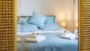 Una cama bien hecha con cubrecama y almohadas azules, una mesita de noche con lámpara y un espejo que refleja la habitación.