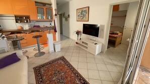 A modern kitchen with orange cabinets, a dining area with bar stools, a flat-screen TV, and a patterned rug. - Exclusive Apartment Sun and Sea  (Novalja)