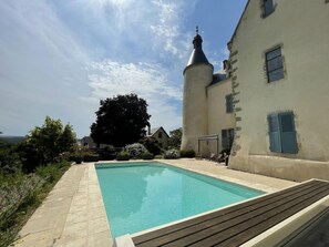 A swimming pool area with a wooden deck, a large tree, and a building with a tower and shutters.